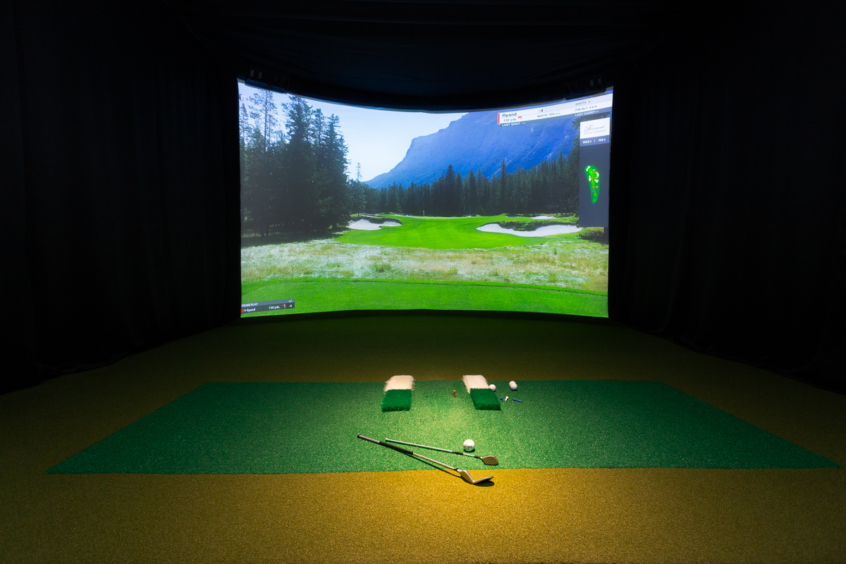 Golf clubs on the hitting mat facing a Banff-style mountain course on the simulator screen